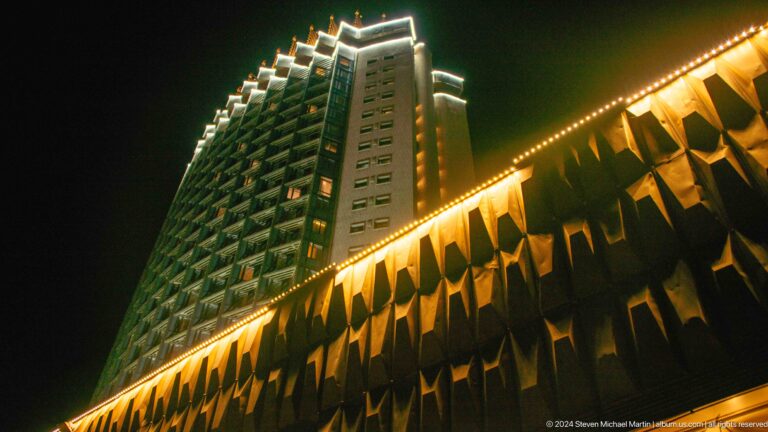Upward night view of a glowing high-rise hotel tower with a patterned, gold-lit podium in Almaty, Kazakhstan.