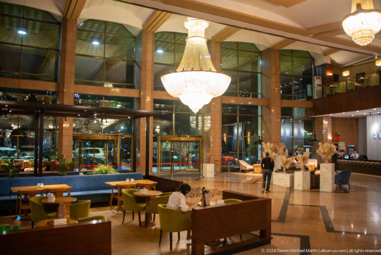Spacious hotel lobby in Almaty with high ceiling, chandeliers, and seating areas arranged around small tables.