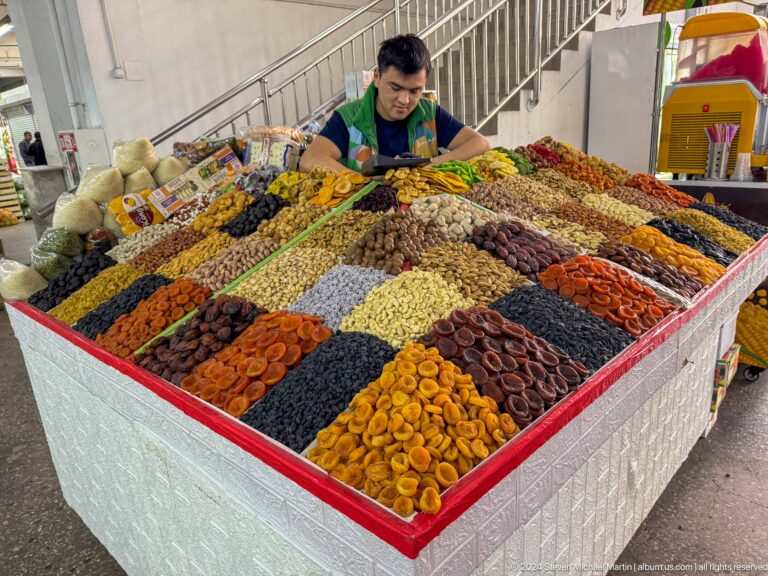 Vendor arranging large bins of neatly ordered dried fruits and nuts at a Green Bazaar stall in Almaty, Kazakhstan.