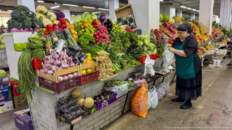 Vendor standing beside a stall in Almaty’s Green Bazaar, with piles of fresh vegetables and herbs displayed on the counter indoors.