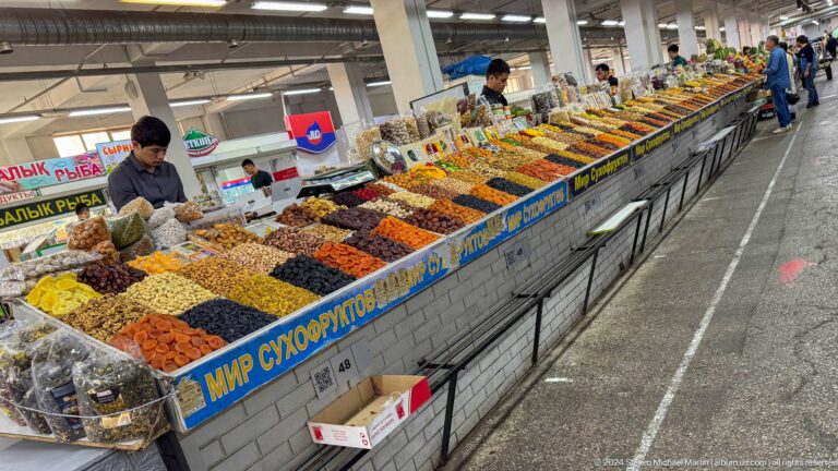 Indoor aisle at Almaty’s Green Bazaar with a long counter displaying parallel rows of dried foods beneath Cyrillic signage.