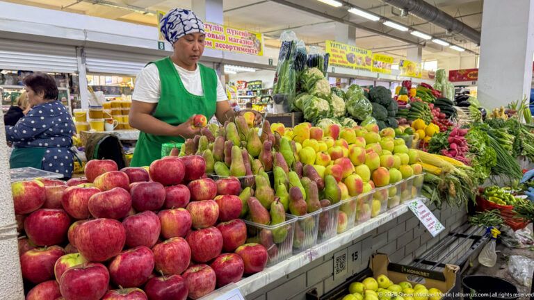 Fruit vendor in an Almaty market standing behind neatly arranged piles of apples and pears.