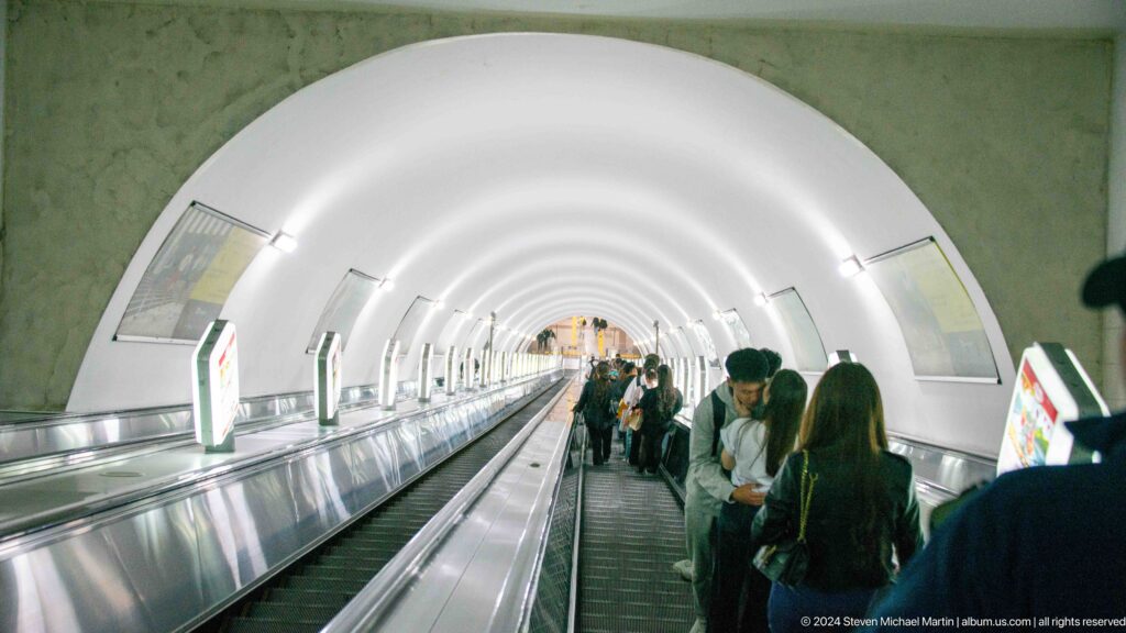 Passengers on the Almaty Metro escalator beneath a smooth white arched tunnel interior.