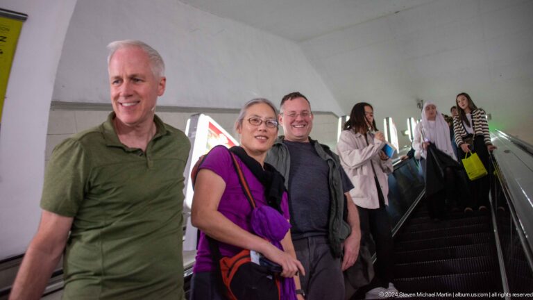 Man and woman smiling at the camera while riding an Almaty Metro escalator, with a white vaulted ceiling and other passengers in the background.