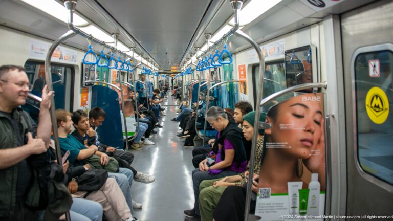 Crowded interior of an Almaty Metro train with passengers seated along both sides of the carriage.