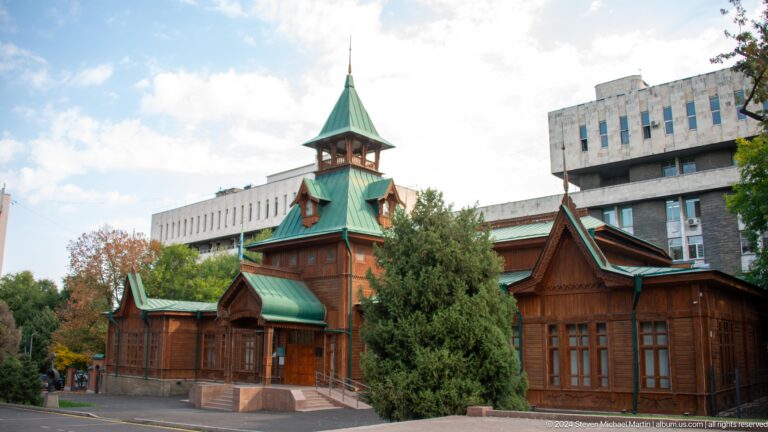 Exterior view of Ascension Cathedral, a wooden Orthodox church with green roofs surrounded by trees in Panfilov Park, Almaty.