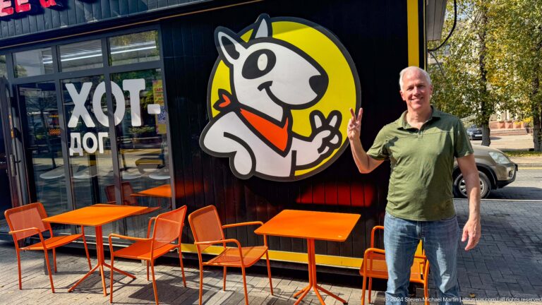 Man standing beside a cartoon sheep logo and orange café chairs at a Hot Dog venue in Almaty, Kazakhstan.