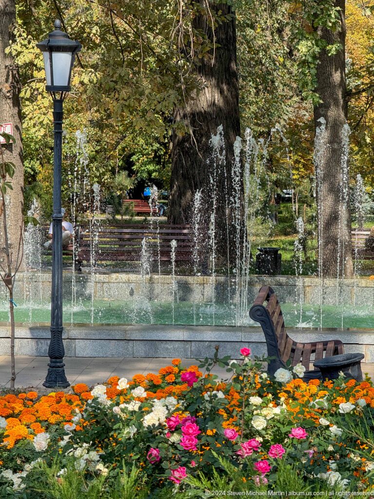 Side view of a landscaped park fountain with vertical water jets, a bench, and flowerbed in an urban park in Almaty, Kazakhstan.