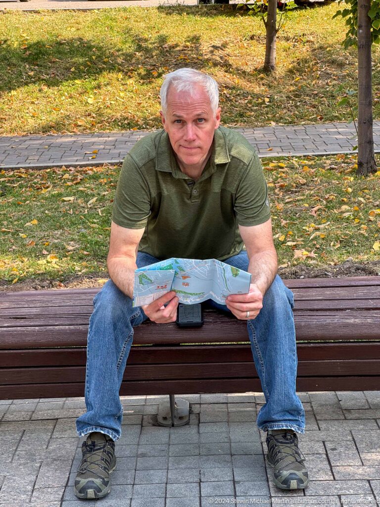 Man seated on a park bench in Almaty reading a large folded city map, with fallen leaves on the ground.