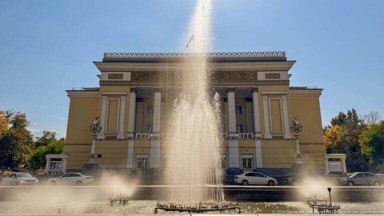 Tall central fountain jet in front of Abai Opera House in Almaty, a pale neoclassical theater with columns and a pediment.
