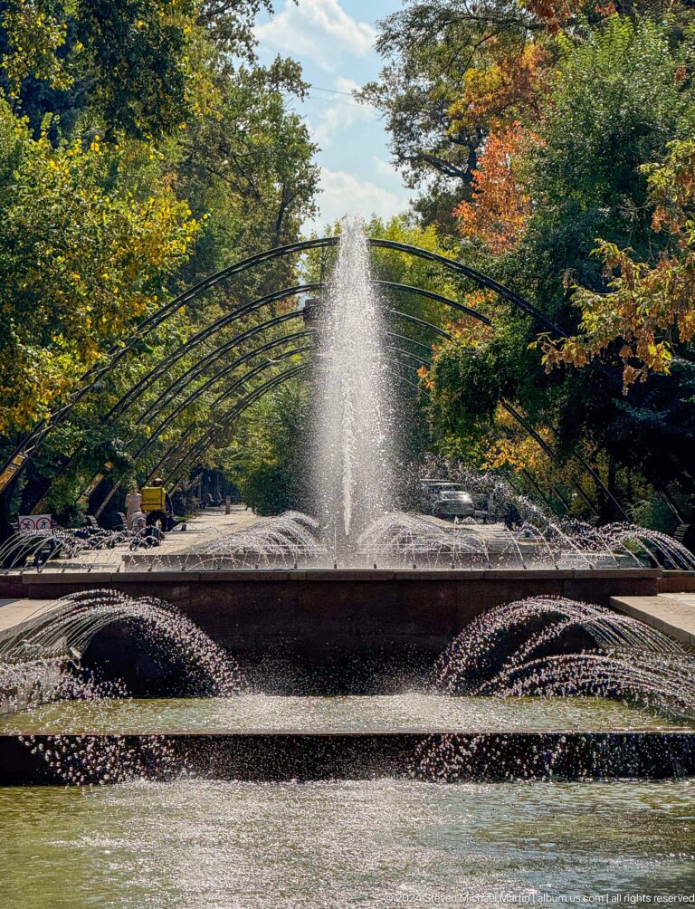 Arched streams of water from a public fountain spray beneath a metal trellis structure in a park setting.