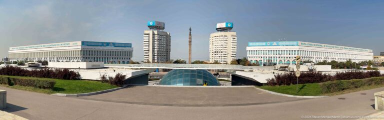 Panoramic view along the axis of Republic Square in Almaty, featuring a central glass dome and a tall monument framed by surrounding urban buildings and open paving.