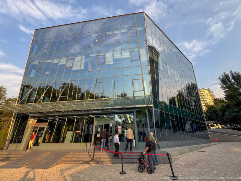 Street-level view of a modern glass commercial building in Almaty with pedestrians near the entrance.