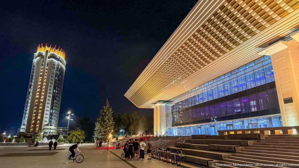 Night view of a central Almaty plaza with a brightly lit cultural building and a tall illuminated tower behind, seen from the front steps with a few pedestrians gathered.