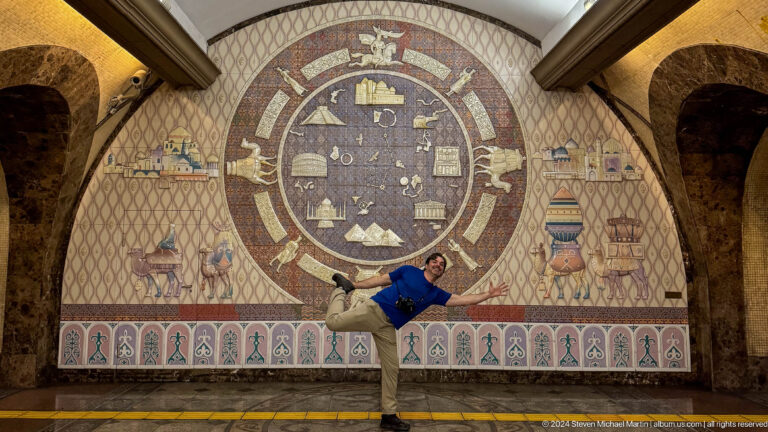 Man posing with extended arms and one leg raised in front of a large circular cultural mural inside the Almaty metro in Kazakhstan.