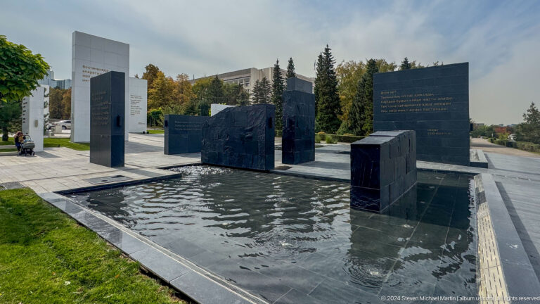 Black stone memorial slabs emerging from a shallow reflecting pool in an urban park setting in Almaty.