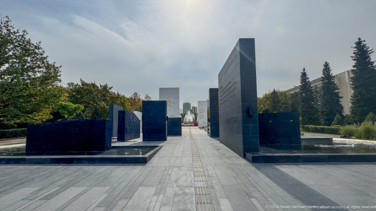 Perspective view down a long paved ceremonial walkway between black memorial walls in an outdoor memorial landscape in Almaty, Kazakhstan.