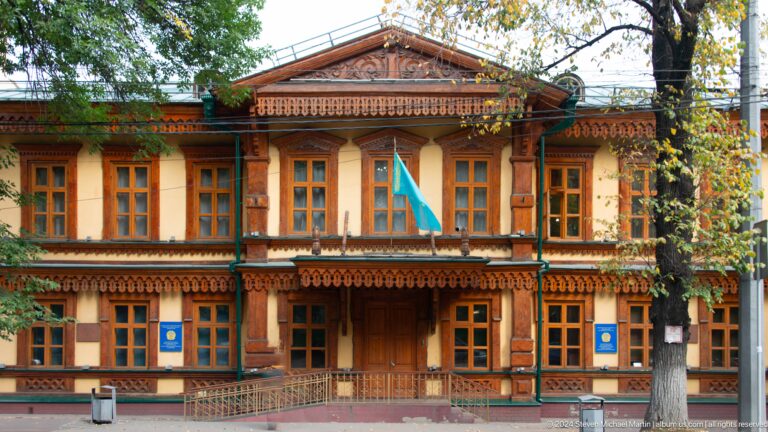 Ornate two-story wooden building in Almaty with carved balconies and decorative wooden detailing on the façade.