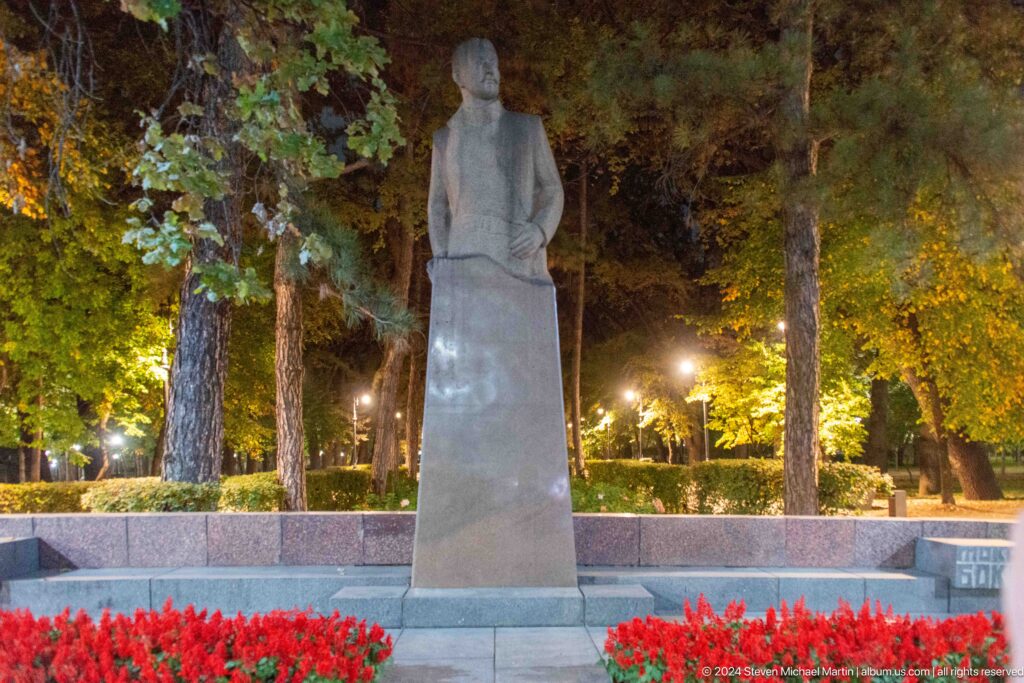 Stone statue on a tall pedestal among trees and red flowers in a park in Almaty, Kazakhstan.