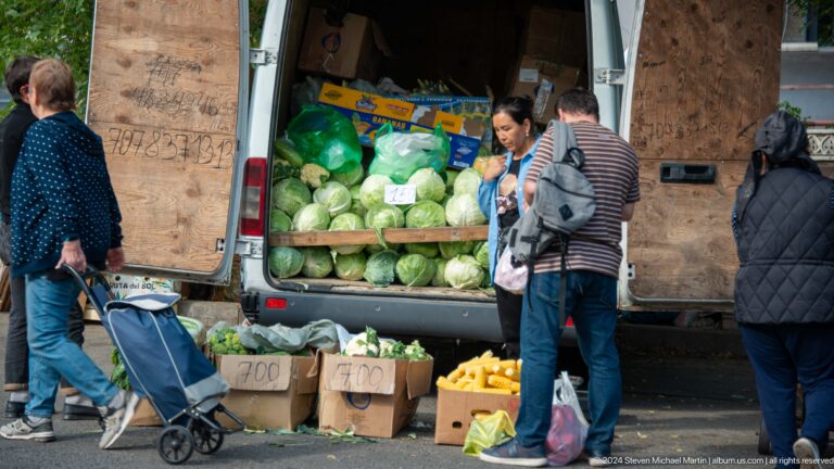 Outdoor vegetable stall in Almaty with cabbages stacked in an open van and corn displayed in boxes in front, with a shopper nearby.