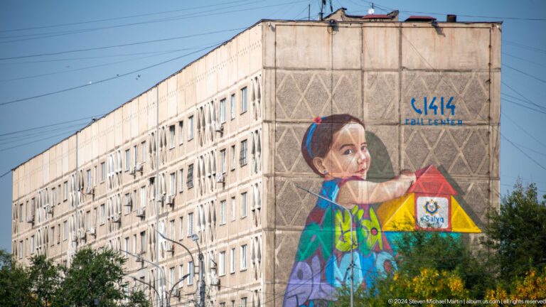 Large mural of a child holding a yellow cup painted on the side of an apartment building in a city street.