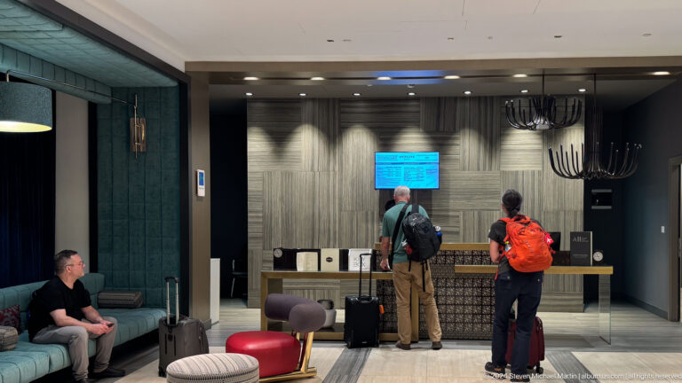 Travelers with backpacks and wheeled suitcases stand at a hotel reception desk in Almaty, Kazakhstan.