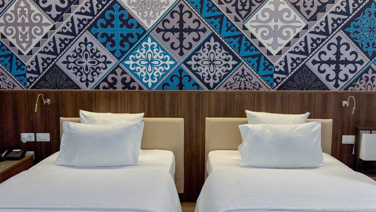 Hotel guest room in Almaty with two single beds beneath a large blue Kazakh-inspired geometric textile panel.