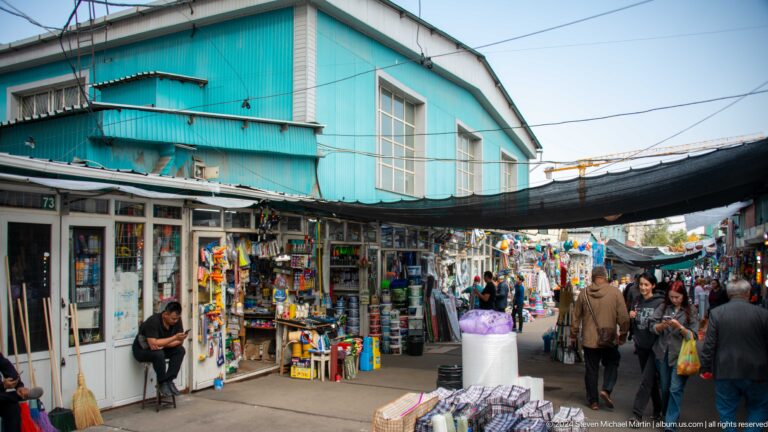 Crowded covered market street in Almaty with a turquoise building above and stalls lining both sides of the walkway.