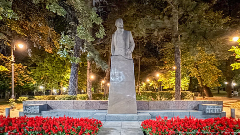 Night view of a park statue standing in a flowerbed beneath tall trees in an Almaty public space.