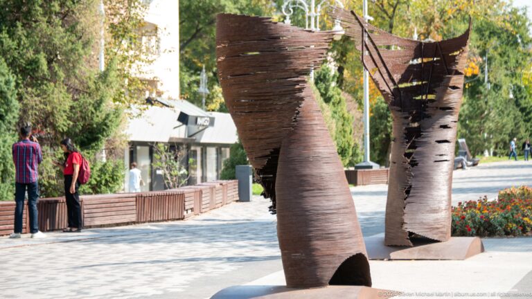 Two tall, twisting abstract metal sculptures stand on a paved plaza with buildings and trees in the background.