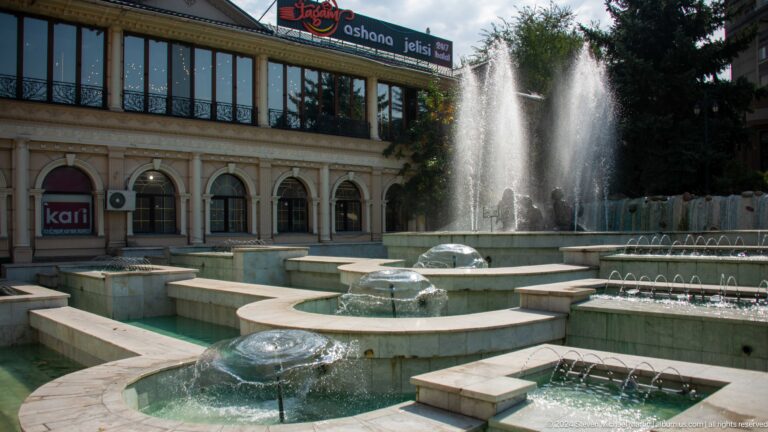 Tiered public fountain basins set in a paved plaza in front of a large civic building with repeated arches.