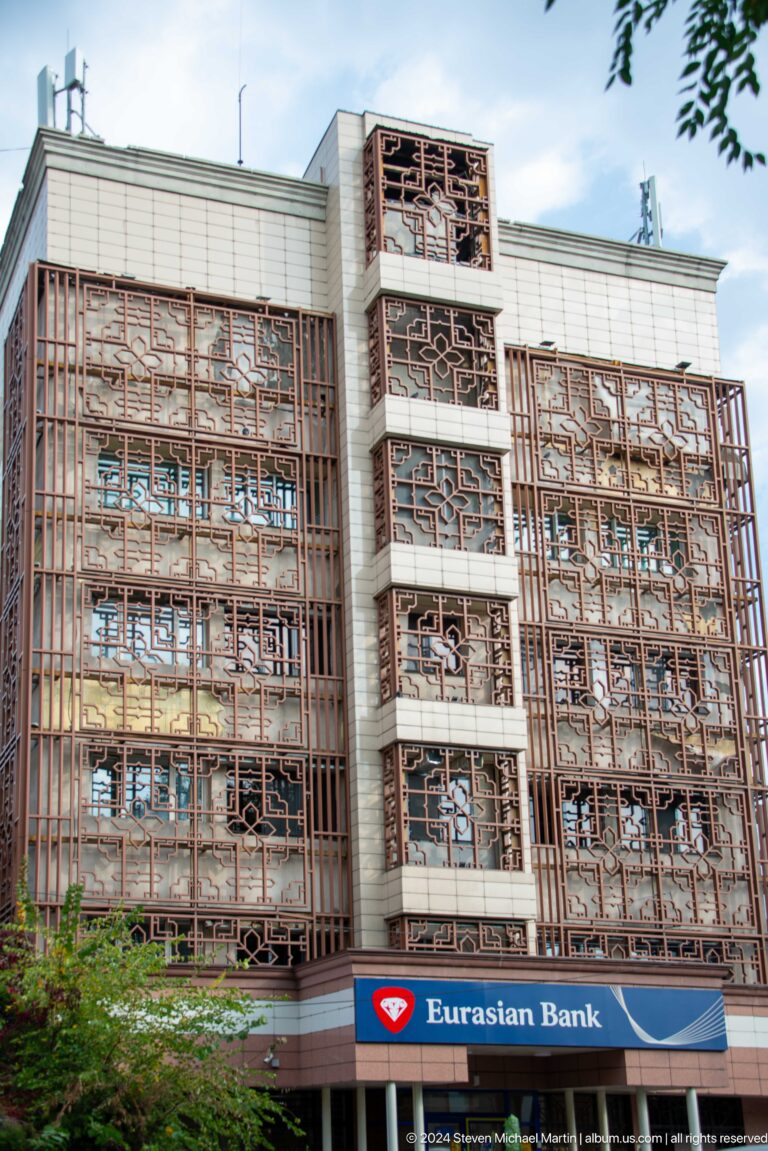 Multi-story modern facade in Almaty wrapped in dense brown metal lattice with white vertical bays.