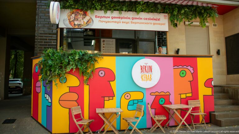 Colorful mural with stylized figures painted on an outdoor café barrier along a city street.