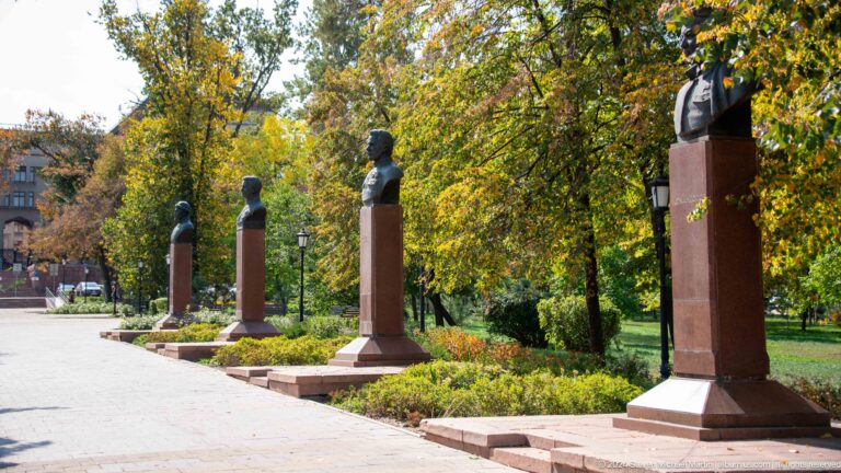 Park walkway in Almaty, Kazakhstan, lined with portrait busts on matching red-granite pedestals among trees and flowerbeds.