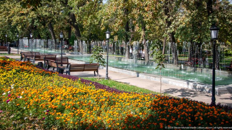Long rectangular fountain bordered by colorful flowerbeds and walkways in an Almaty urban park.