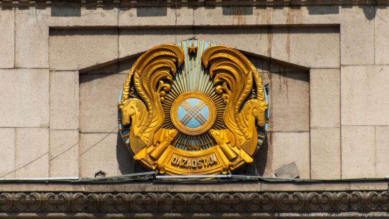Gold Kazakhstan coat of arms mounted on a light stone building facade.