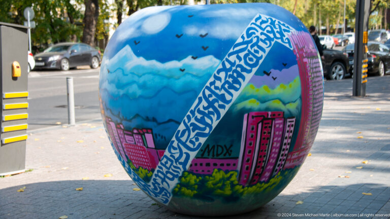 Painted spherical sculpture in an Almaty public space, decorated with clouds and stylized city buildings.