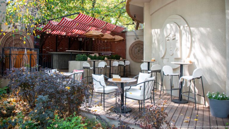 Outdoor café terrace in Almaty with seating beneath a large red awning along a city street.