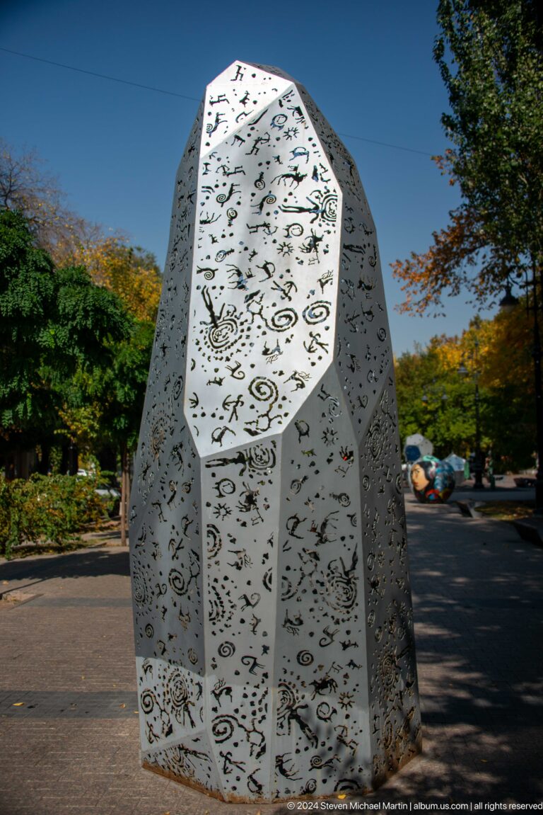 Tall perforated metal column with abstract decorative patterns standing outdoors as a public sculpture.
