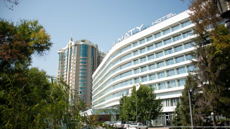 Curving white modern hotel facade with adjoining tower against a clear sky in Almaty.