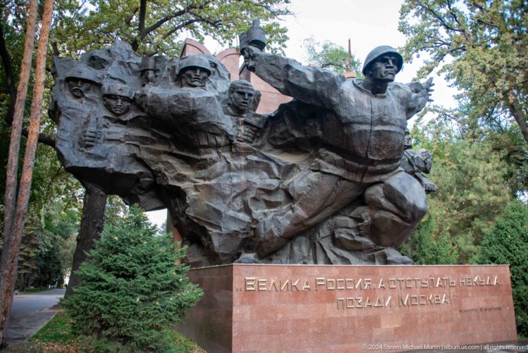 Detail of the Panfilov heroes bronze war memorial in Almaty, Kazakhstan, showing helmeted Soviet soldiers emerging from a rough sculptural mass above a red stone base.