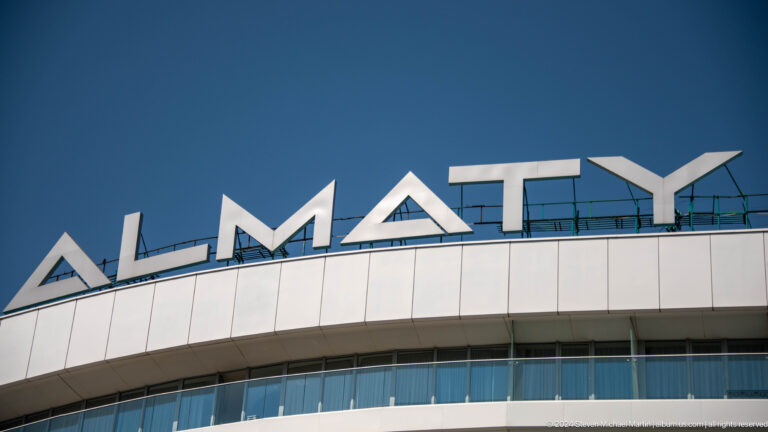 Large white ALMATY letters on the roof of a modern curved building against a clear blue sky.