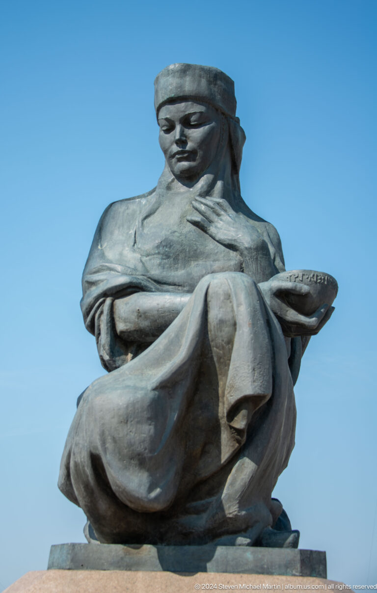 Close-up of a seated bronze statue of an intellectual figure in Almaty, Kazakhstan, shown in draped clothing with one hand on the chest and the other holding a rounded inscribed object.