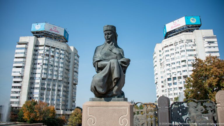 Full view of a bronze statue of a seated historical figure on a stone pedestal in an outdoor public space.