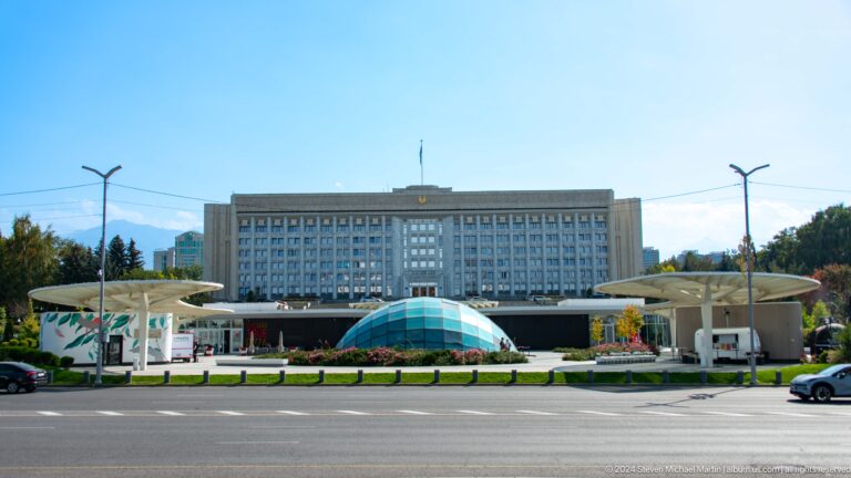 Government building at Republic Square in Almaty, Kazakhstan, seen across a plaza with a low blue-domed fountain.