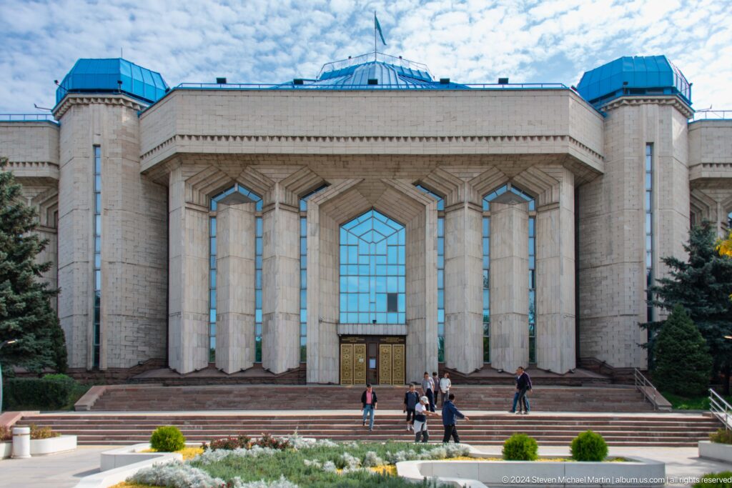 Wide view of the Central State Museum of Kazakhstan exterior with tall columns, a blue central window, and steps leading up from a paved plaza.