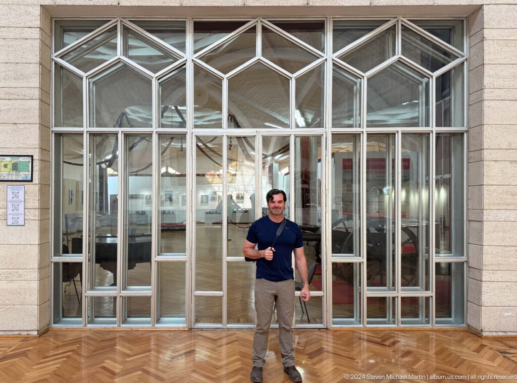 Medium view of a modern visitor posing at the glass entrance doors of the Central State Museum in Almaty, Kazakhstan.