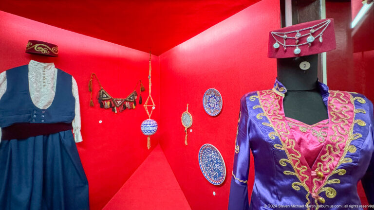 Wide view of a traditional costume with jewelry displayed against a red exhibition backing in the Central State Museum in Almaty, Kazakhstan.