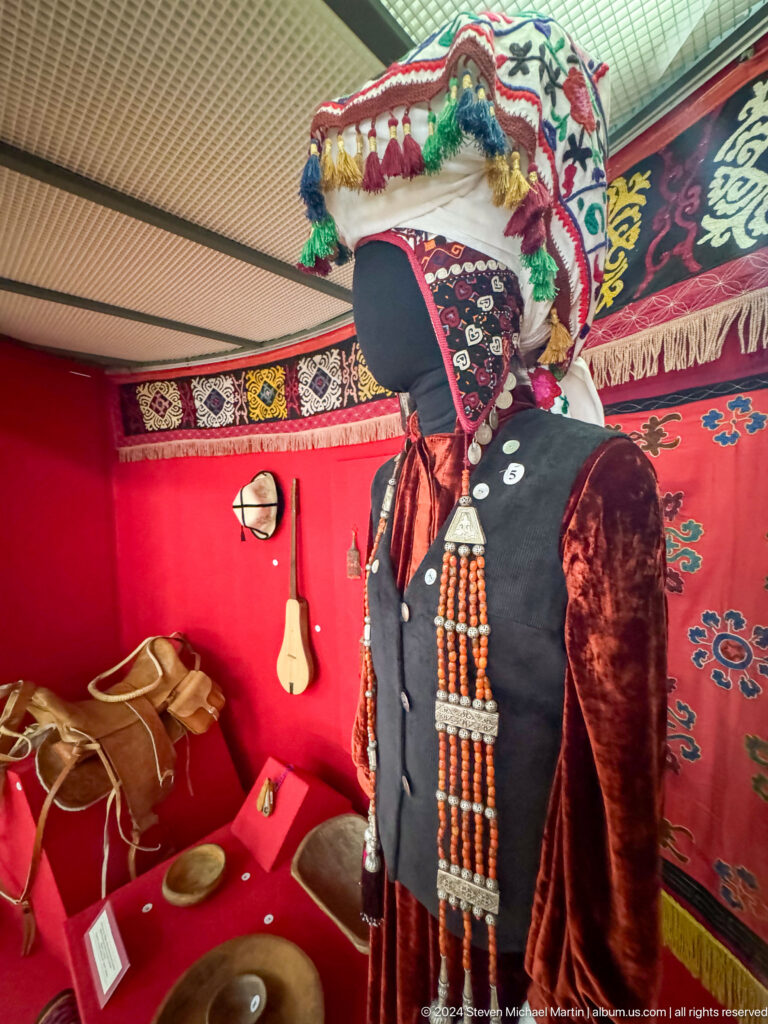 Traditional embroidered coat, hat, belt, and small accessories mounted against a red display backing in the Central State Museum in Almaty, Kazakhstan.