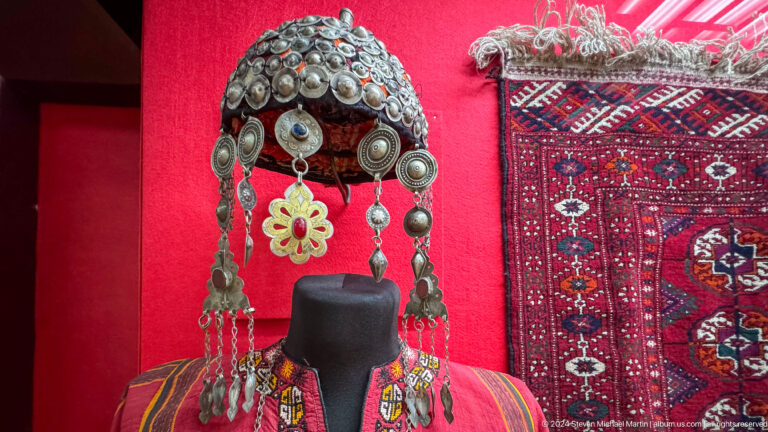 Ceremonial silver headdress with hanging ornaments displayed beside patterned textile in the Central State Museum of Kazakhstan.